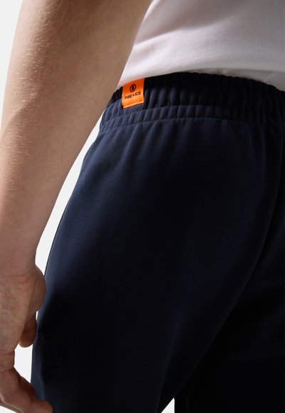 Zbliżenie na osobę w ciemnoniebieskich spodniach z elastycznym pasem, z pomarańczową etykietą „FIRE + ICE” oraz białą koszulą wpuszczoną do spodni.