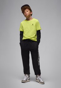 T-shirt amarelo néon com logótipo preto, usada sobre uma camisa preta de mangas compridas; calças de treino pretas com o texto branco "JORDAN". Ténis cinzentos e brancos.