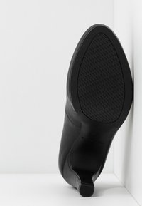 Chaussure à talon haut noire positionnée à la verticale contre un mur d'angle blanc, montrant la semelle texturée et le talon fin.