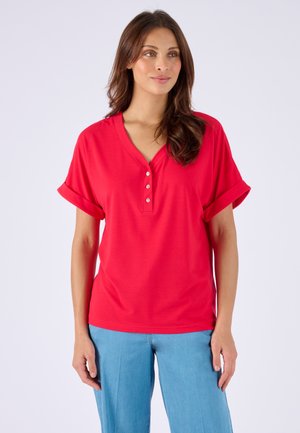 Donna che indossa una maglietta henley rossa a maniche corte con polsini arrotolati e pantaloni azzurri, in piedi contro uno sfondo semplice.