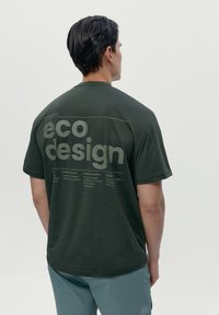 Temno zelena majica s sproščenim krojem, ki prikazuje besedilo "eko oblikovanje" v beli barvi, ter oznake s podrobnostmi o načelih trajnosti na hrbtni strani.