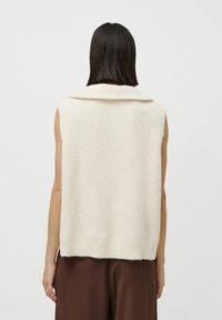 Persoană cu păr drept și închis la culoare poartă un top din tricot crem fără mâneci și pantaloni maro, întorcându-se cu fața de la un fundal alb simplu.