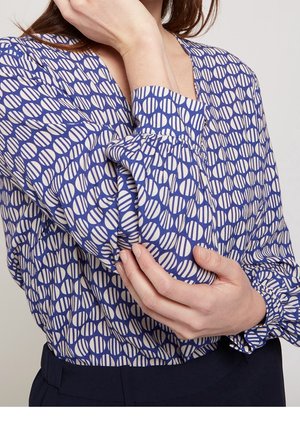 Kvinde iført blå- og hvidmønstret bluse med lange ærmer og rynkede manchetter, holder sin modsatte arm nær albuen.