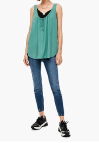 Camisa sin mangas verde claro con escote fruncido y detalle de cordón, combinada con jeans oscuros ajustados y zapatillas negras.