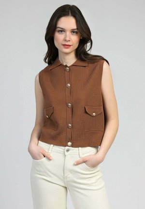 Femme aux cheveux longs et foncés portant un top marron sans manches avec des boutons et un col, ainsi qu'un pantalon couleur crème, les mains dans les poches, fond neutre.