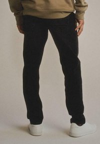 Persona che indossa jeans neri, sneakers bianche e una camicia a maniche lunghe marrone, vista di spalle su uno sfondo semplice.