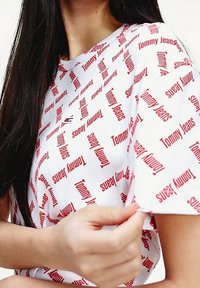 Valkoinen puuvillapaita, jossa on punainen "Tommy Jeans" -teksti eri suuntiin, miehustassa pyöreä kaula-aukko ja lyhyet hihat.