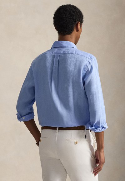 Ανοιχτό μπλε λινό πουκάμισο με γυρισμένα μανίκια, γιακά και λεπτομέρεια πτυχής στην πλάτη. Συνδυάζεται με κρεμ παντελόνια που διαθέτουν διακοσμητικό ράψιμο.