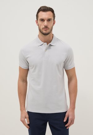 Uomo con capelli corti castani e barba indossa una polo a maniche corte grigio chiaro e pantaloni scuri, in piedi davanti a uno sfondo chiaro uniforme.