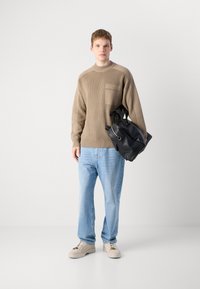 Young man standing wearing beige sweater, light blue jeans, beige sneakers, holding black leather backpack against white background.