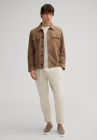 Braune Wildlederjacke mit zwei Brusttaschen, Knopfverschluss und leichtem Material, getragen über einem hellen Pullover und cremefarbenen Hosen.