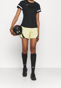 Camiseta deportiva negra de Nike con mangas cortas blancas, pantalones cortos amarillos a rayas horizontales, calcetines negros y un balón de fútbol negro.