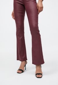 Benen met glanzende kastanjebruine wijde broekspijpen en zwarte open-toe hoge hak sandalen, hand rustend langs het dijbeen op een witte achtergrond.