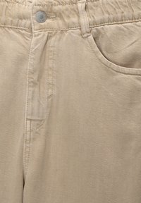Pantalones de algodón beige con cinturilla elástica, cierre con botón y bolsillos laterales. La tela tiene una sutil textura diagonal.