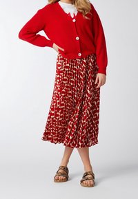 Czerwony sweter z dzianiny z dużymi guzikami, w połączeniu z spódnicą midi z geometricznym wzorem w kolorach czerwonym i kremowym. Brązowe sandały z klamrami.