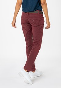 Burgundy slim-fit pants with subtle back pockets, textured belt, and a smooth cotton finish. Worn with white athletic shoes.