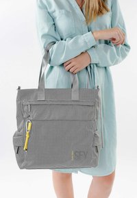 Frau in hellblauem Kleid hält eine große graue Tragetasche mit vorderer Reißverschlusstasche und gelbem Reißverschlusszug.