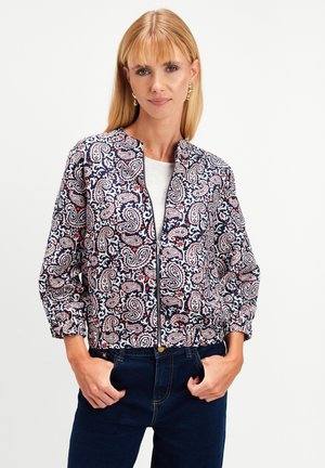 Blondynka w złotych kolczykach w kształcie kropli, zapinanej kurtce z wzorem paisley w niebieskim, różowym i białym kolorze, białej koszuli i ciemnoniebieskich dżinsach.