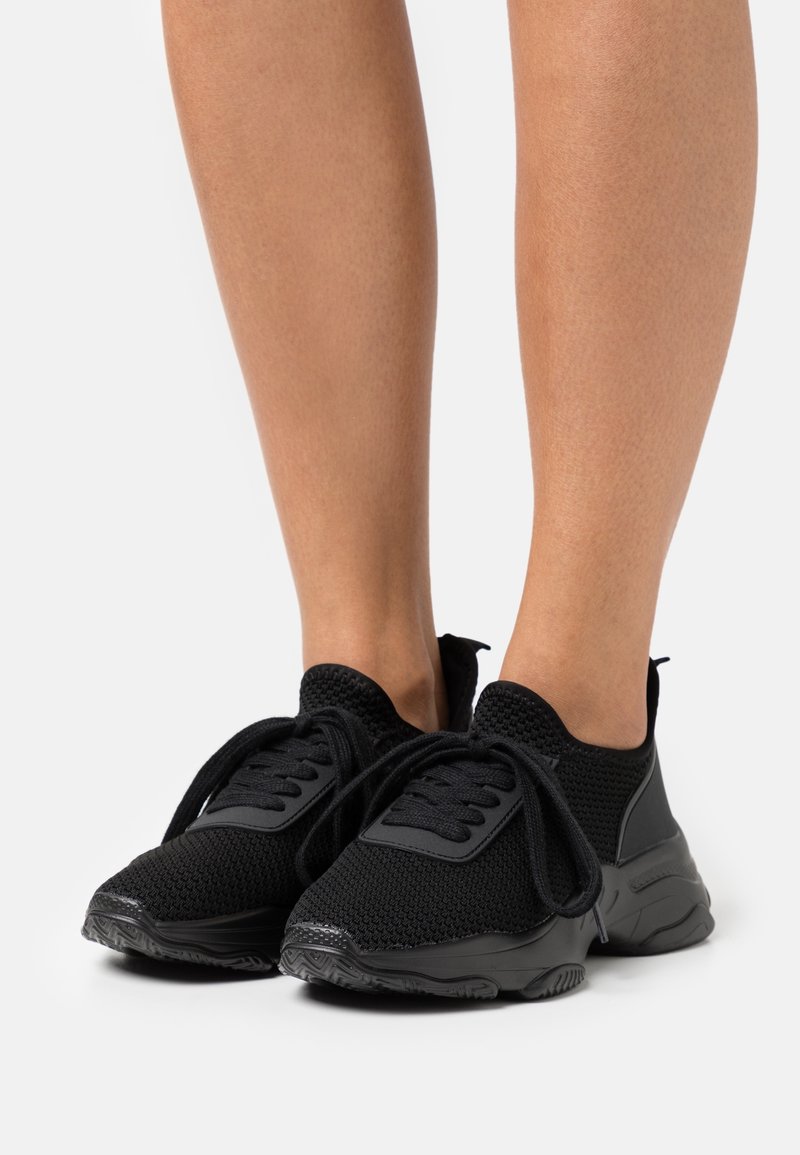 Jambes portant des baskets de sport en maille noire avec des semelles épaisses et des lacets, montrées sur un fond clair uni.