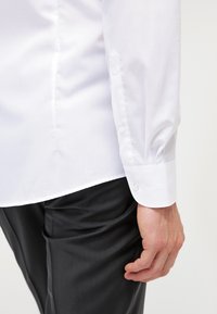 Chemise blanche à manches longues avec un col structuré, des poignets boutonnés et un tissu lisse et léger, associée à un pantalon foncé et brillant.