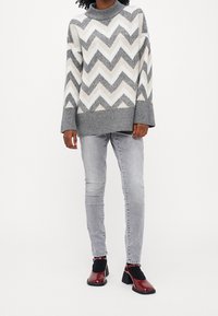 Grobstrickpullover mit grau-weißer Zickzackmuster, hohem Kragen, kombiniert mit hellgrauen Jeans und bordeauxfarbenen Blockabsatzschuhen.