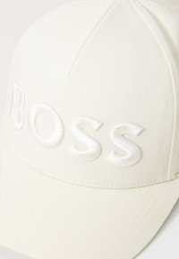 Hvid kasket lavet af tekstureret stof med en broderet "BOSS"-logo i hvid. Designet inkluderer en buet skygge og syet detaljer.