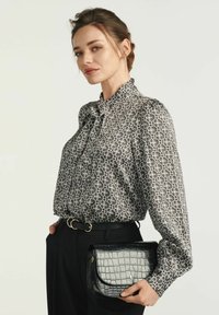 Czarna i biała bluzka w wzory z wysokim dekoltem i detalem w formie kokardy, zestawiona z czarną torebką o tłoczeniu w kształcie krokodyla oraz czarnymi spodniami.