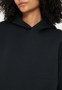 Femme aux cheveux bouclés portant un sweat à capuche noir uni, lèvres fermées, visage partiellement visible du nez vers le bas.