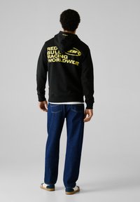 Sudadera negra con el texto amarillo "Red Bull Racing Worldwide", combinada con jeans de mezclilla azul. Presenta una capucha y un corte estándar.