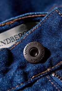 Μπλε υφαντό τζιν με χρυσή ραφή, με μεταλλικό κουμπί σκούρου χρώματος με λογότυπο και ορατή ετικέτα. Κοντινή λήψη που δείχνει την υφή και τις λεπτομέρειες.