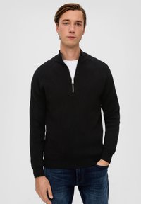 Schwarzer gerippter Pullover mit einem Reißverschluss-Kragen, der lange Ärmel und eine figurbetonte Silhouette hat. Über einem weißen T-Shirt getragen.