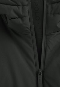 Svart jacka med hög krage, mjuk textur och framdragkedja med "POLO CLUB" präglat på tejpen runt dragkedjan.