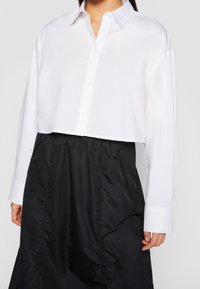 Chemise blanche courte à boutons avec col classique, assortie à une jupe noire texturée dotée de poches zippées.