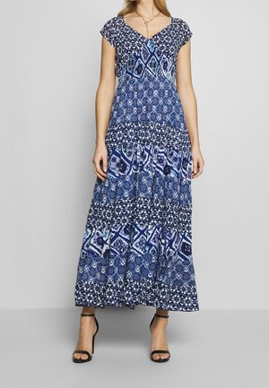 Woman wearing a blue patterned maxi dress with short sleeves and v-neck, paired with black ankle-strap high-heeled sandals.