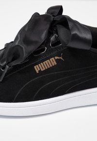 Baskets en daim noir Puma avec de larges lacets en ruban de satin et une semelle blanche texturée, affichant le logo Puma doré sur le côté.