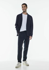 Marinblå kavaj med strukturerade axlar, vit t-shirt, skräddarsydda marinblå byxor och vita sneakers med grå accenter. Slätt tyg, avslappnad passform.