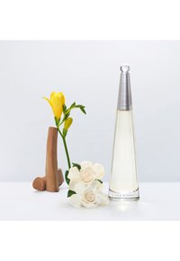 Botella de vidrio transparente en forma de cono con una tapa plateada, rodeada de flores de rosa blancas y una flor amarilla en un jarrón de madera.