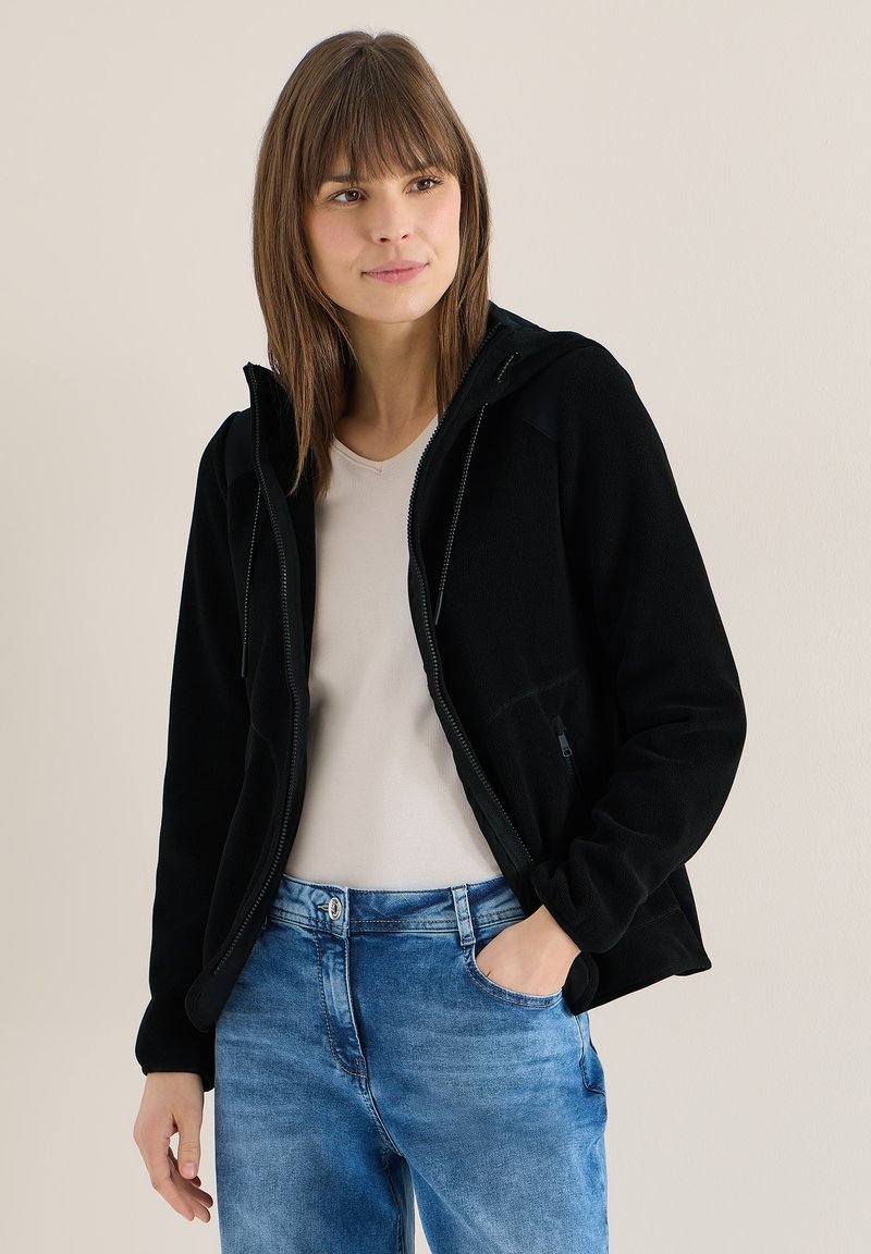 Femme portant une veste polaire noire à fermeture éclair sur une chemise beige clair et un jean bleu, une main dans la poche, debout devant un fond uni.