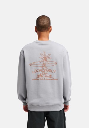 Homme portant un sweat-shirt gris clair avec un motif marron de palmier et de planche de surf et le texte "Locals Only" dans le dos.