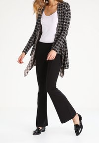 Kvinna som bär svarta utsvängda byxor, vit ärmlös topp, svart mönstrad öppen kardiagan och svarta glänsande loafers som går på en vit bakgrund.