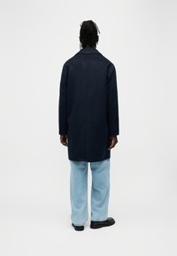 Manteau bleu marine à silhouette longue, col cranté et fente simple au dos. Porté avec un jean bleu clair et des chaussures noires, en tissu texturé.