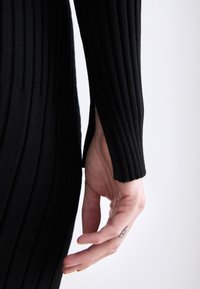 Black ribbed knit dress sleeve, featuring a side slit and textured pattern. Hand shown with a tattoo and slightly curved fingers.