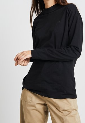 Persona que lleva una camisa negra de manga larga y pantalones beige, de pie frente a un fondo claro y liso con las manos entrelazadas.