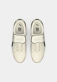 Hvide sneakers med sorte detaljer, med en velcro-strop, tekstureret overdel, snørebånd og gummisål. Inkluderer mærkets logo på stroppen.