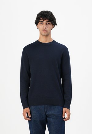 Ung mand med kort krøllet sort hår iført en enkel mørk marineblå sweater og blå jeans mod en hvid baggrund.