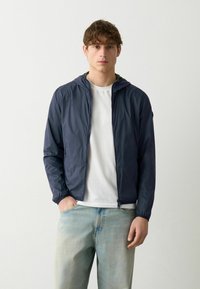 Marinefarbene leichte Jacke mit Kapuze, durchgehendem Reißverschluss und elastischen Bündchen, kombiniert mit hellblauen Jeans und einem weißen T-Shirt.