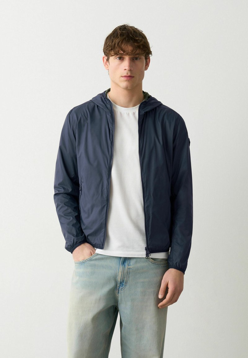 Marinefarbene leichte Jacke mit Kapuze, durchgehendem Reißverschluss und elastischen Bündchen, kombiniert mit hellblauen Jeans und einem weißen T-Shirt.