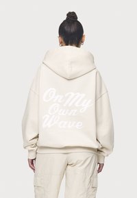 Beige oversized hoodie med en huva och stor vit text på baksidan som säger "On My Own Wave." Mjuk material och lös passform.
