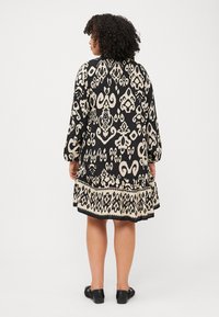 Une personne aux cheveux bouclés tourne le dos, portant une robe à motifs noirs et crème avec des manches longues et un ourlet volanté.