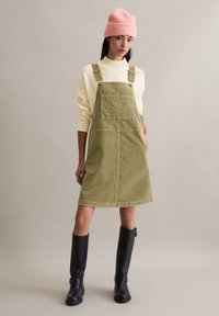 Groene corduroy jurk met een vierkante zak, verstelbare bandjes en zijzakken, gedragen over een crèmekleurige trui en gecombineerd met zwarte laarzen tot over de knie.